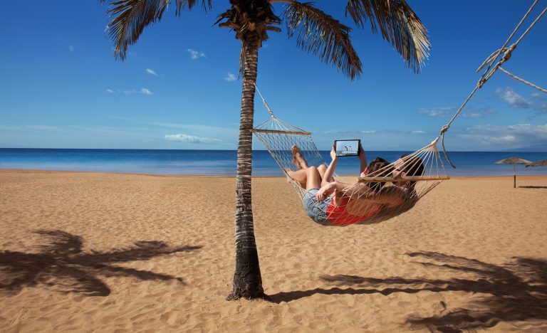 Photo of a couple at the beach, lying in a hammock and looking at a tablet computer