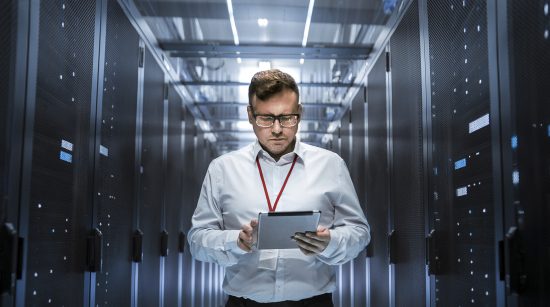 IT Technician Walks Through Rows of Server Racks in Data Centre. Simultaneously He Works on a Tablet Computer.