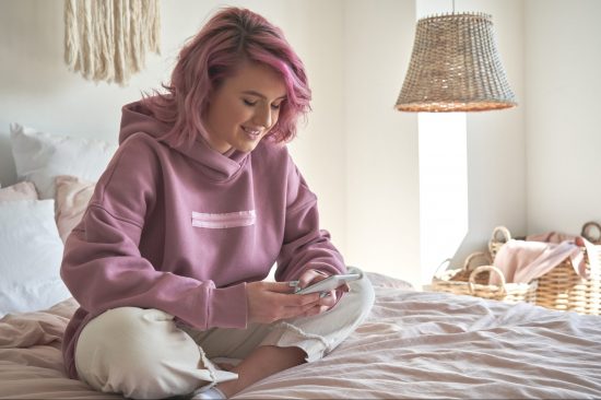 Smiling hipster teen girl with pink hair holding smart phone chatting in social media, watching video content, texting messages, playing games, using mobile apps, surfing internet sit in bed at home.